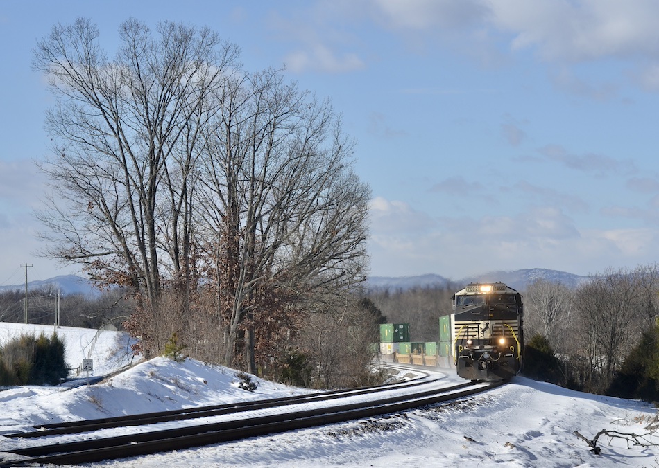 A Norfolk Southern intermodal freight train passes through a snowy landscape.